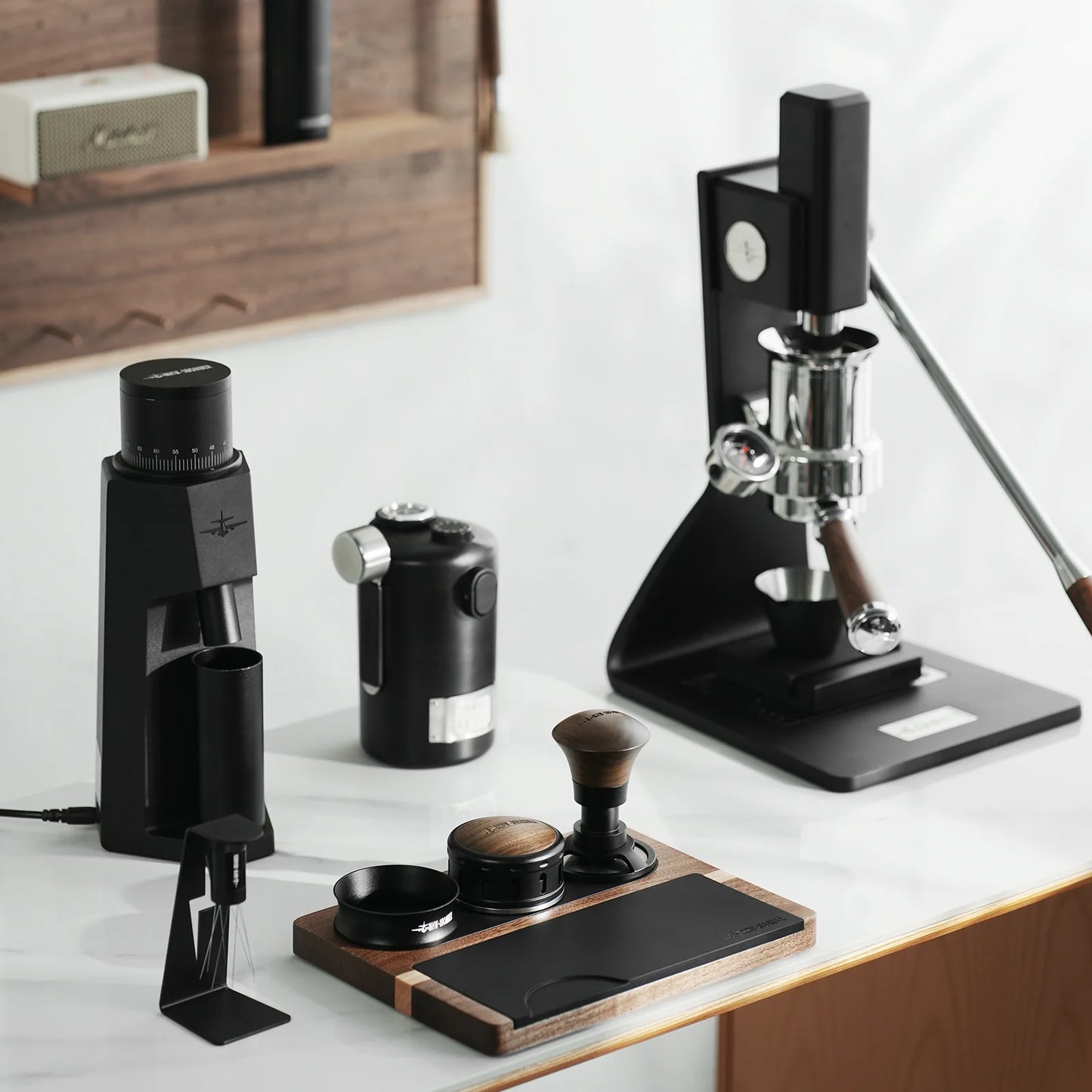 A sleek coffee setup on a white countertop features a modern grinder, kettle, and advanced coffee press with adjustable tamping depth. Surrounded by barista accessories like the MHW-3BOMBER Armor Series Coffee Tamper 2.0, the background includes a wooden shelf with more coffee essentials.