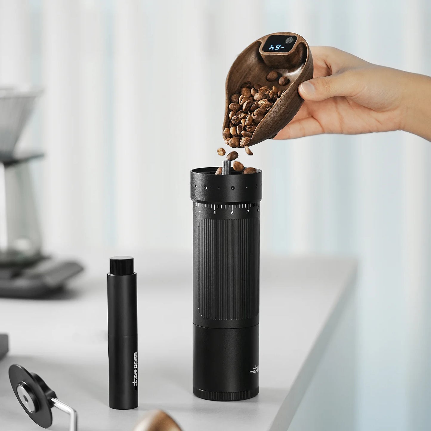 A person pours coffee beans with a wooden scoop into a black grinder on a white countertop, surrounded by scattered parts. Soft natural lighting highlights the MHW-3BOMBER Ovo Coffee Scale & Spray Bottle Set placed to the side, essential for the brewing process.
