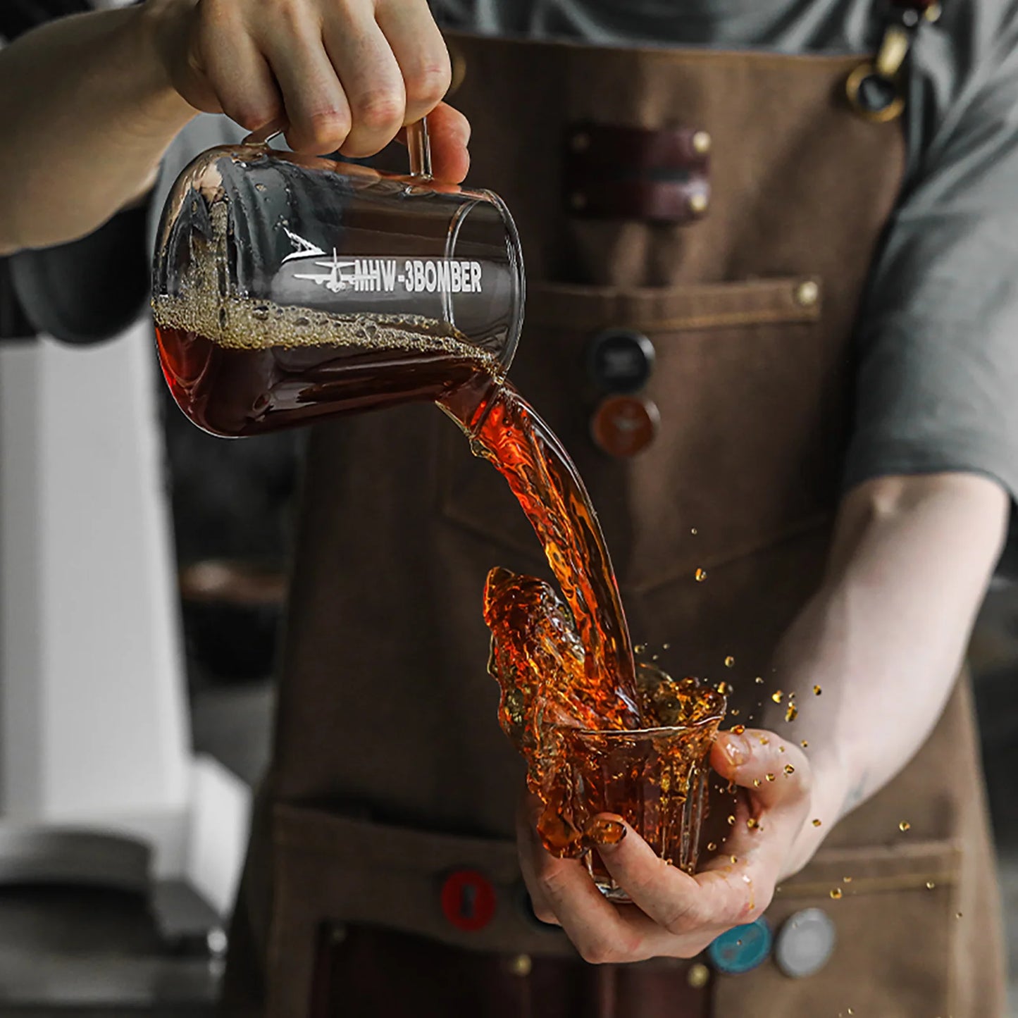 A person in a brown apron skillfully pours a dark liquid, likely coffee, from an MHW-3BOMBER Elf Coffee Server into a small glass cup. The drip-free pour catches the light mid-stream, sending droplets splashing gracefully against the blurred indoor backdrop.