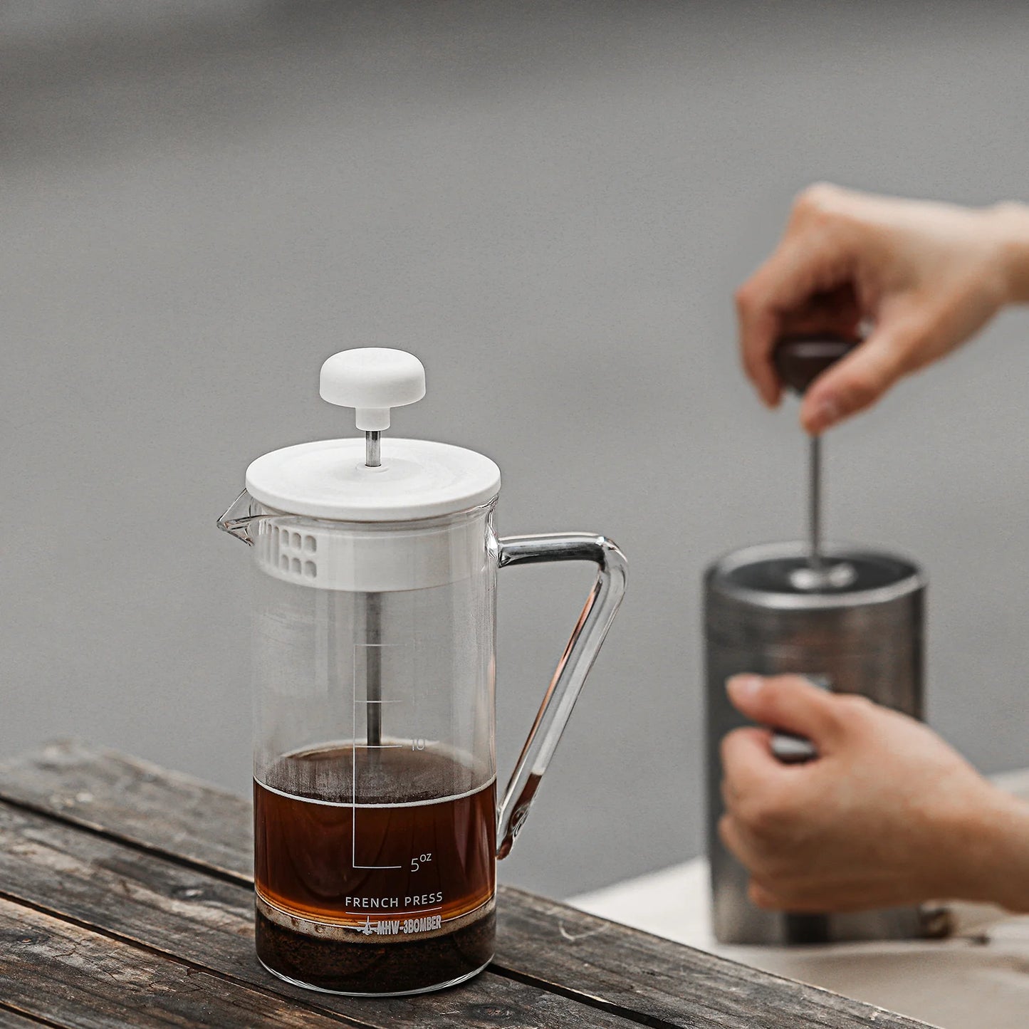 Someone uses a manual coffee grinder next to a MHW-3BOMBER Gimme French Press, featuring a stainless steel filter and partially filled with coffee on a wooden table. Both items have matching white accents, showcasing an elegant setup in heat-resistant glass.