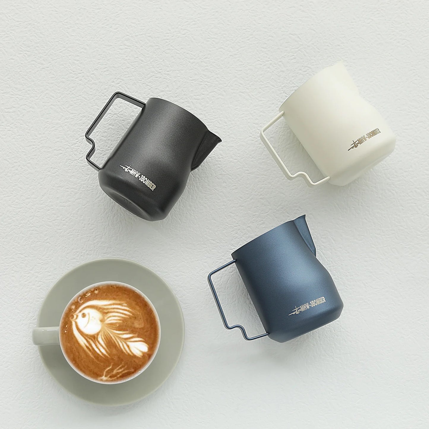 A grey cup with a latte featuring intricate art rests on a light surface, accompanied by three milk frothing pitchers: black, navy blue, and the sleek MHW-3BOMBER Turbo Milk Pitcher in white.