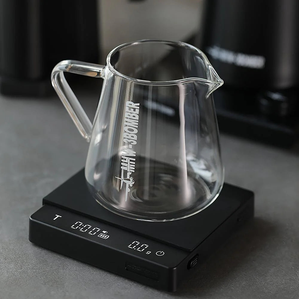 A clear glass pitcher with Tiny Bomber in white rests on the MHW-3BOMBER Espresso Scale Cube Coffee Scale 2.0, offering precise measurements on a digital display. The convenient, rechargeable USB feature enhances this sleek dark setup.