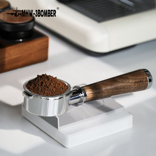 An MHW-3BOMBER Bottomless Portafilter filled with ground coffee is on a white scale in a well-lit room. Its natural walnut wood handle gleams, while a wooden box and stainless steel coffee machine are partially visible in the background.