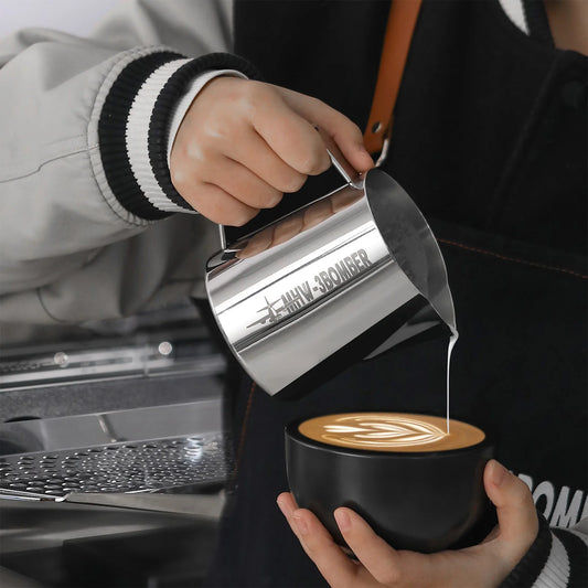 A person in a black and white jacket pours steamed milk from the MHW-3BOMBER Latte Art Pitcher 5.0 Glossy, featuring a distinctive crocodile spout design, into a black cup to create latte art, with an austenitic 304 stainless steel espresso machine gleaming nearby.