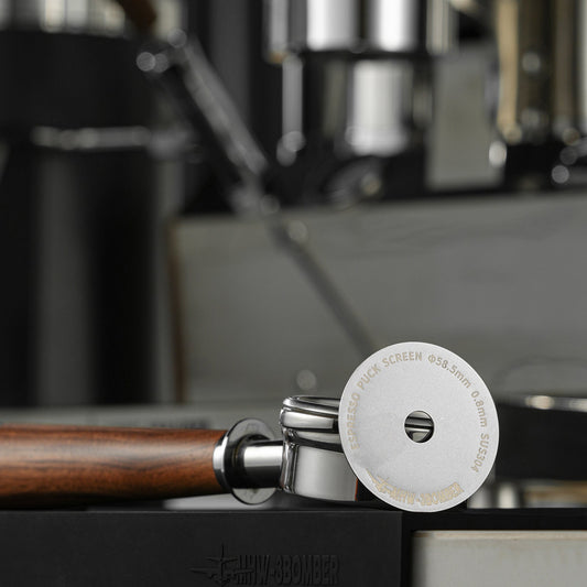 A close-up of the MHW-3BOMBER Espresso Puck Screen with Round Hole, titanium-coated and 58mm, resting on a wooden-handled portafilter. The engraved screen sits on a dark, reflective surface with a blurred background.