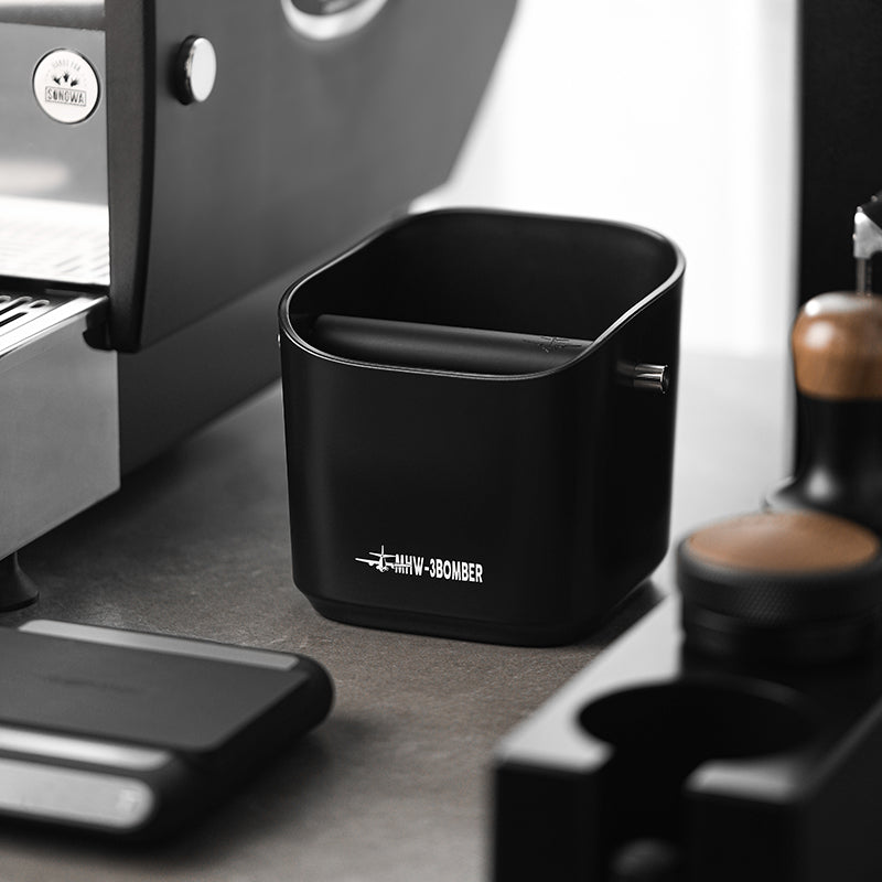 A durable MHW-3BOMBER Coffee Espresso Square Knock Box is on the counter, easy to clean and neatly placed beside an espresso machine and coffee tools like a tamper and small storage container.