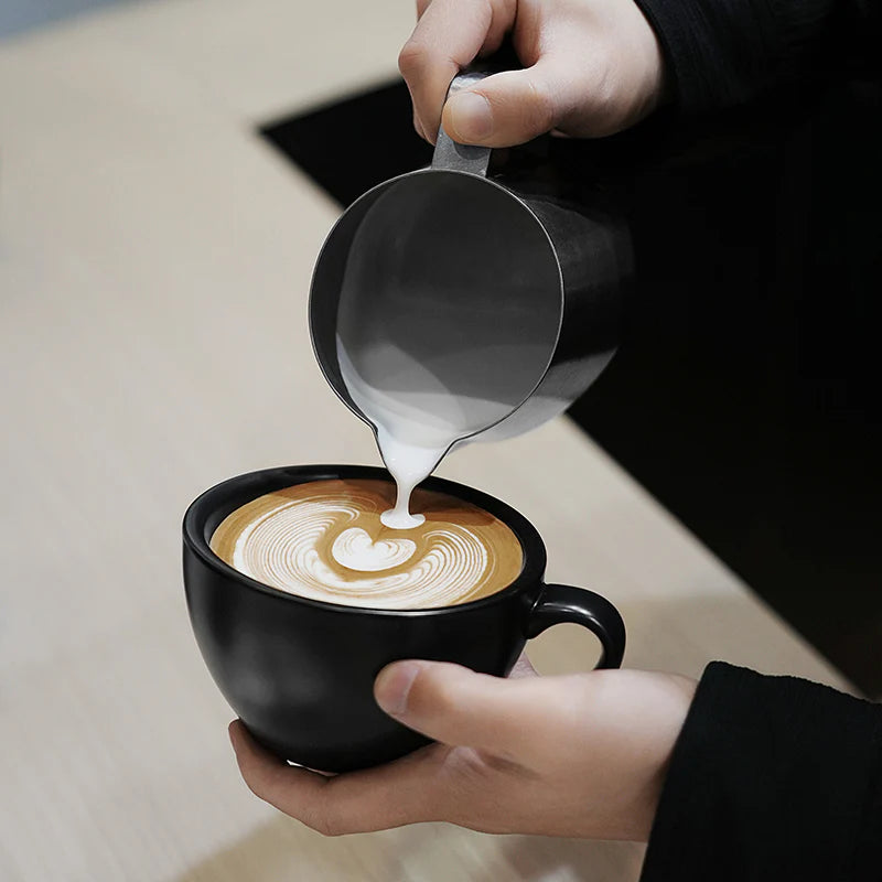 MHW-3BOMBER GT milk pitcher pouring latte art in a black cup. Stainless steel pitcher.