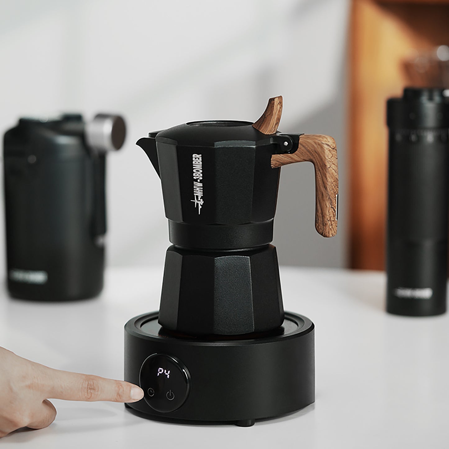 MHW-3BOMBER Electric Coffee Heater. Black Moka pot on base with wood handle, on counter. Finger pressing button.