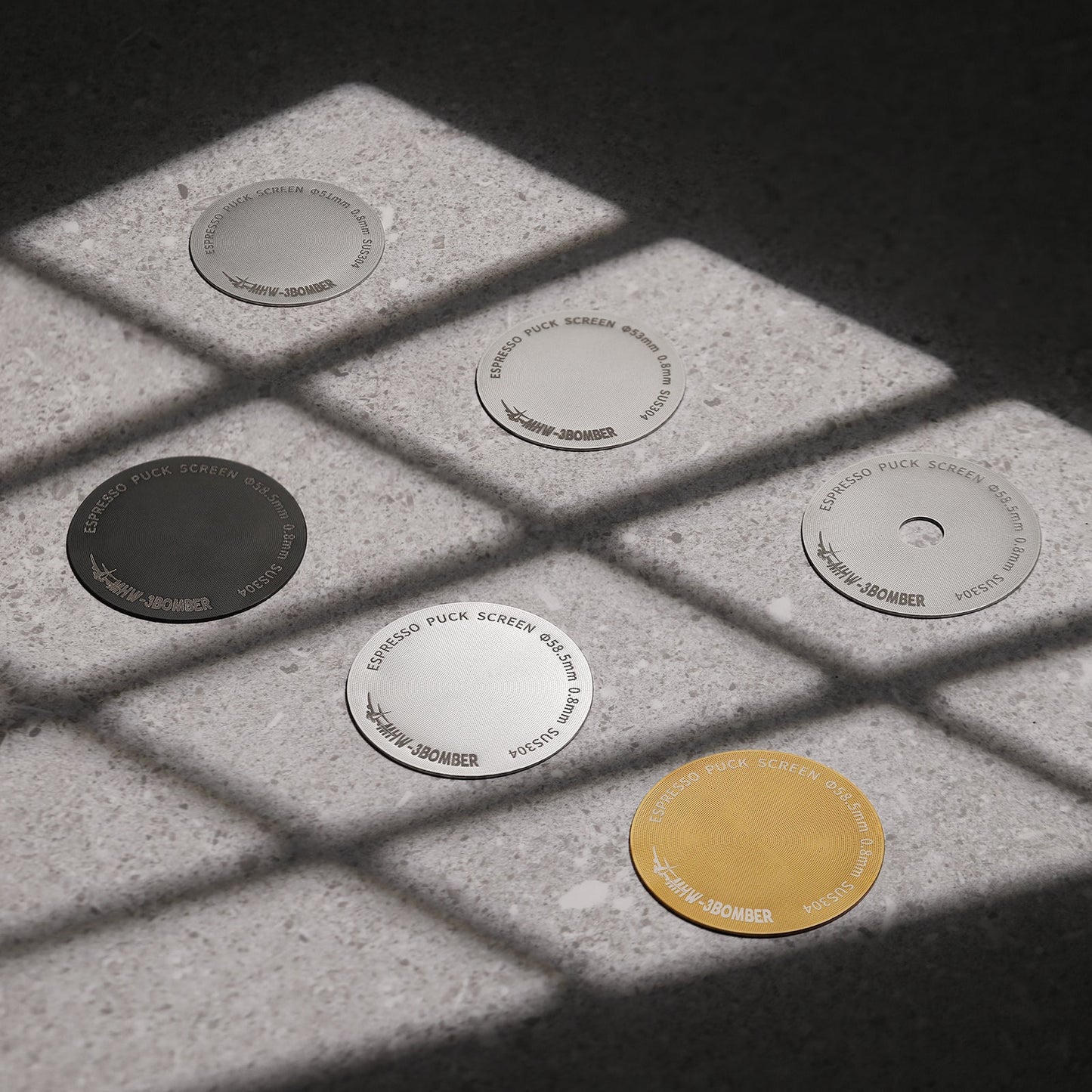Six MHW-3BOMBER Espresso Puck Screens with titanium coating lie on a textured surface, displaying lines of light and shadow. Each disc is inscribed with #38OMBER and comes in finishes like silver, black, or gold.