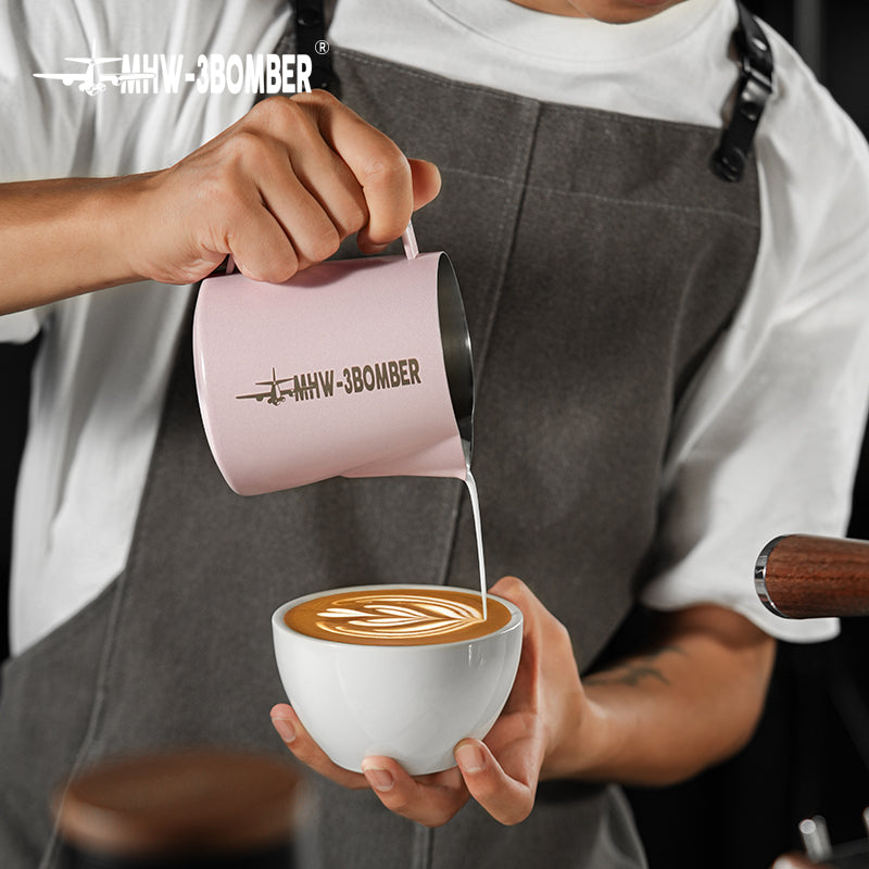 A person wearing a dark apron pours steamed milk from a white MHW-3BOMBER Latte Art Pitcher 5.0 into coffee, using the crocodile spout to create floral latte art.