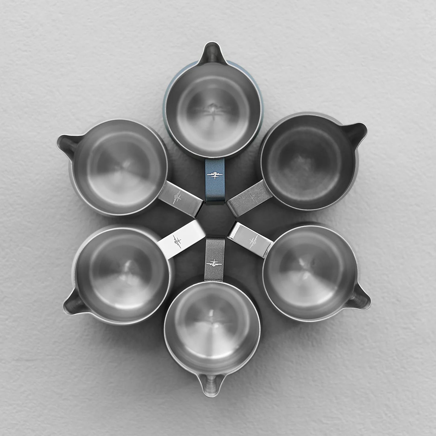 Six MHW-3BOMBER measuring cups, reminiscent of the Turbo Milk Pitcher Glossy, are arranged in a circular, flower-like pattern on a gray surface. The handles radiate outward, creating a symmetrical design similar to graceful milk frothing art.