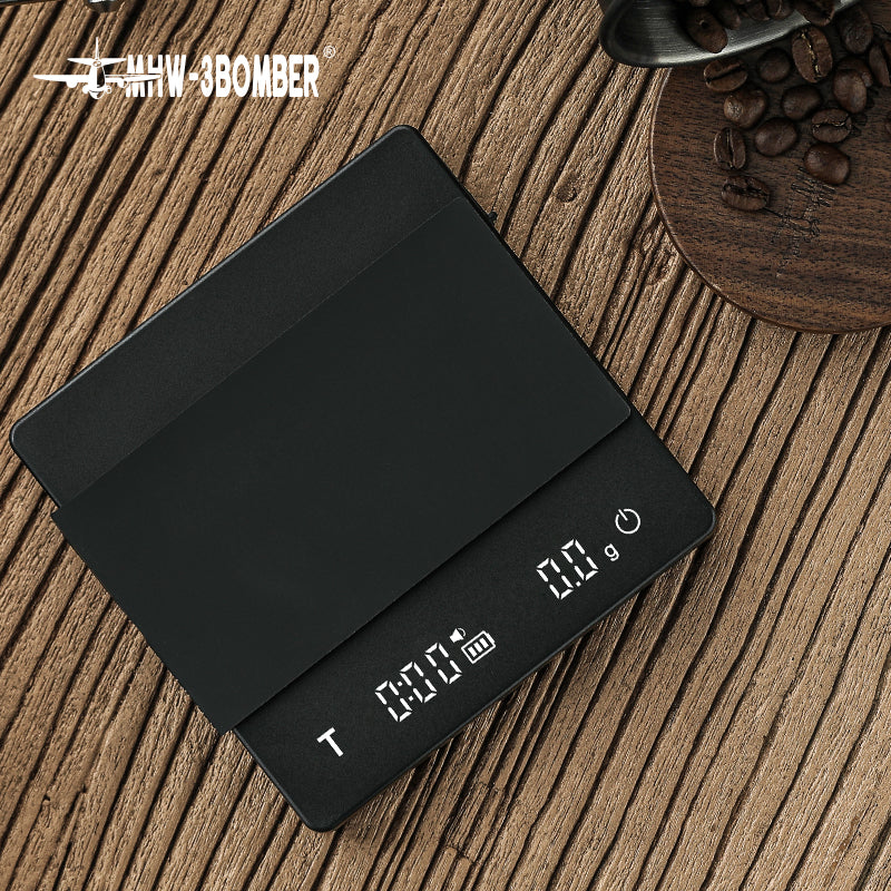 The MHW-3BOMBER Cube Coffee Scale 2.0 Mini, a sleek black digital scale, USB rechargeable and highly precise, rests on a wooden surface. Its LED display shows zero next to a bowl of coffee beans, making it an ideal tool for coffee enthusiasts.