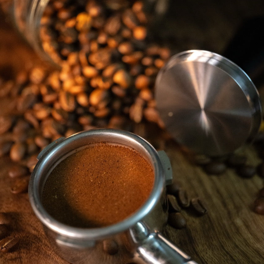 FIX COFFEE tamper with grounds, stainless steel, spring mechanism visible, loose coffee beans on wood.