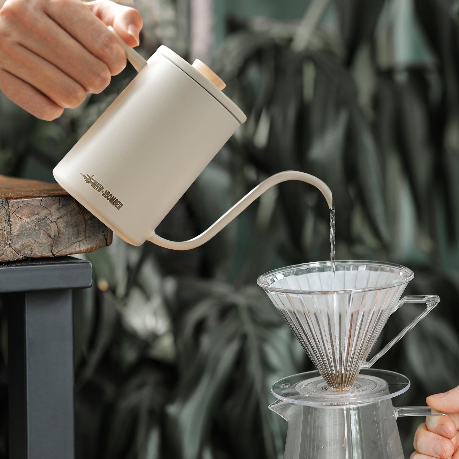 An MHW-3BOMBER Planet Hand Brewing Kettle in pale beige elegantly pours hot water into a glass pour-over coffee dripper on a transparent carafe, set against lush green foliage for an ideal brew setting.