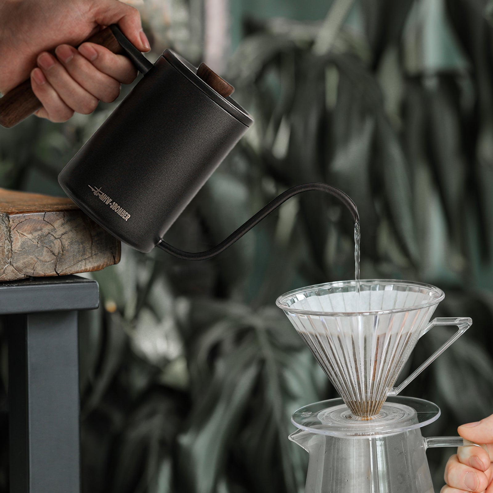 The MHW-3BOMBER Planet Hand Brewing Kettle, with a wooden handle, elegantly pours hot water into a glass pour-over dripper on a stand, against a backdrop of lush green plants.