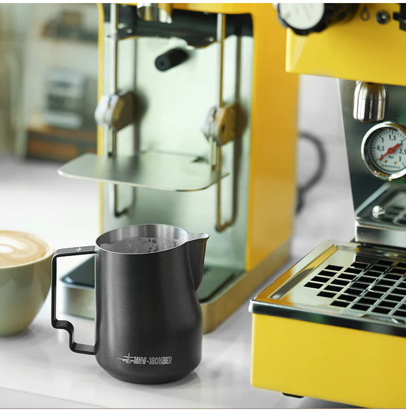 A MHW-3BOMBER Turbo Milk Pitcher Matte Black sits beside a latte with exquisite art on the countertop. In the background, a yellow espresso machine with a pressure gauge and drip tray enhances the modern kitchen, perfect for mastering milk frothing techniques.