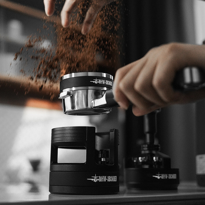 MHW-3BOMBER portafilter holder, black, for espresso tamping, on counter with coffee grounds being poured in.