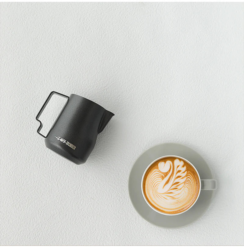 Aerial view of a latte with intricate art in a gray saucer, paired with the MHW-3BOMBER Turbo Milk Pitcher Matte Black, set against a light textured background.