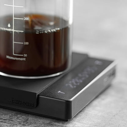 A clear glass beaker with dark liquid rests on the MHW-3BOMBER Espresso Scale Cube Coffee Scale 2.0, which offers precise digital readings and USB rechargeability. The sleek design and textured gray surface enhance this modern setups elegance.