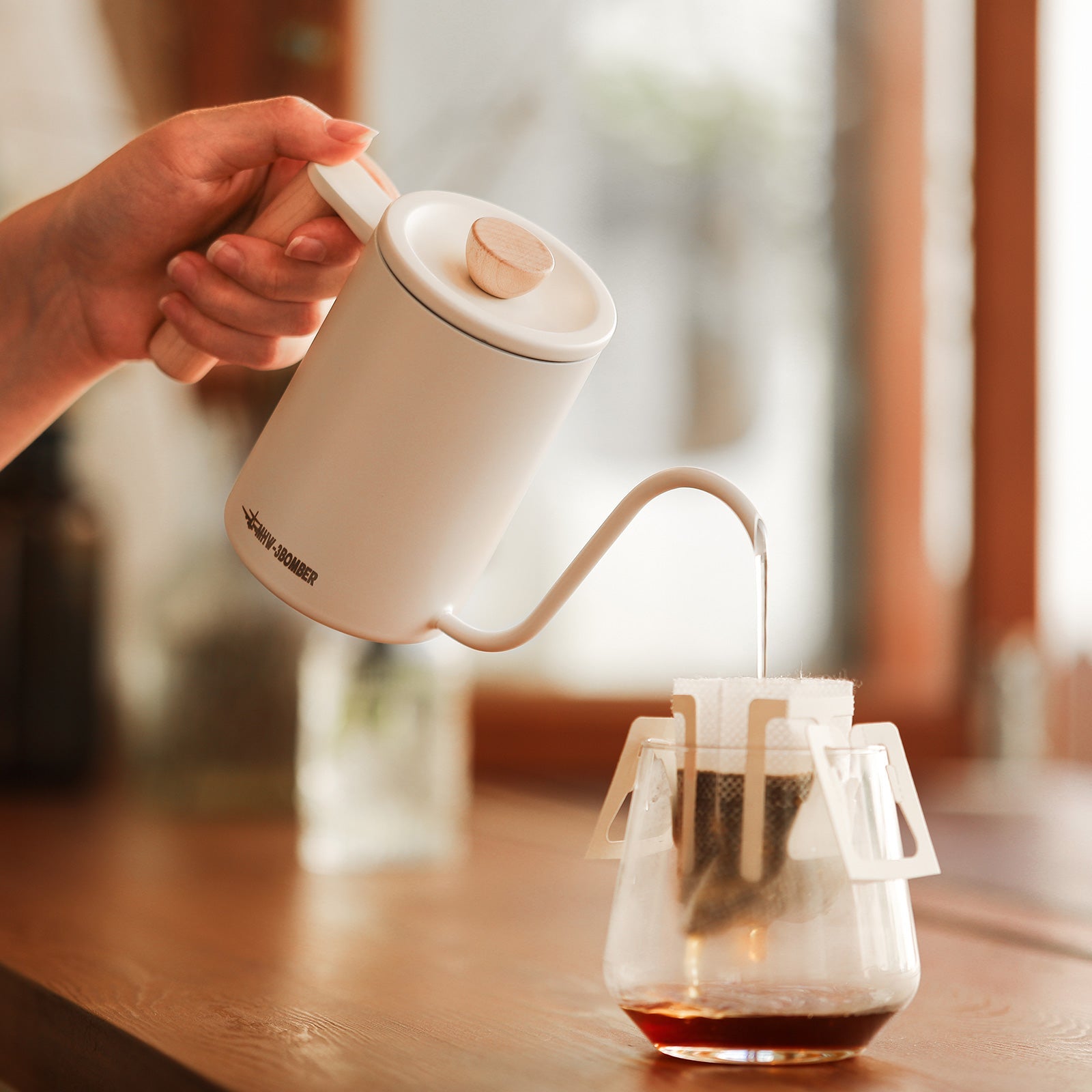 A hand pours steaming water from a MHW-3BOMBER Planet Hand Brewing Kettle into a drip coffee filter over a glass carafe, with rich coffee slowly brewing at the bottom against a softly blurred backdrop of wooden and glass elements.