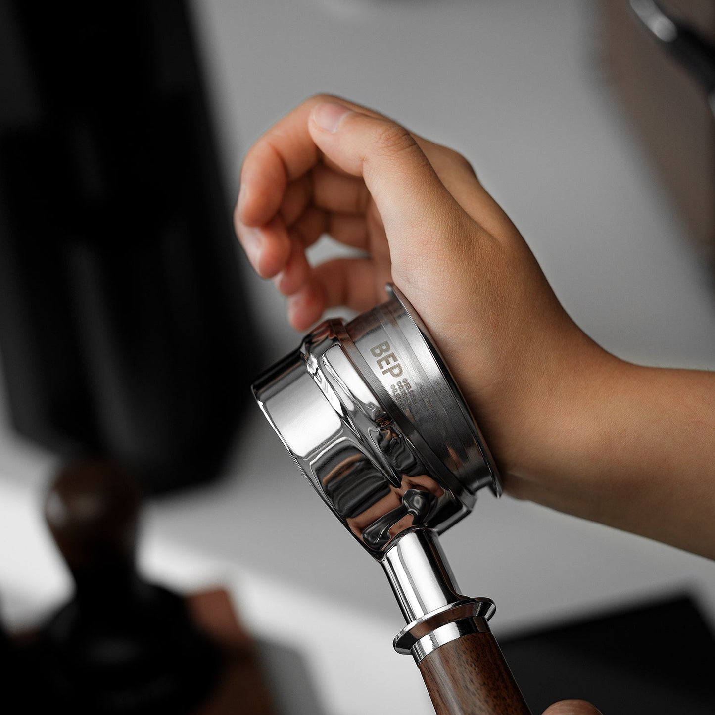 A hand holds the MHW-3BOMBER BEP Butterfly Coffee Filter Basket 2.0 with a chrome portafilter and wooden handle, highlighting its dual-layer filtration against a blurred background of a tamper and other stainless steel coffee tools on a white surface.