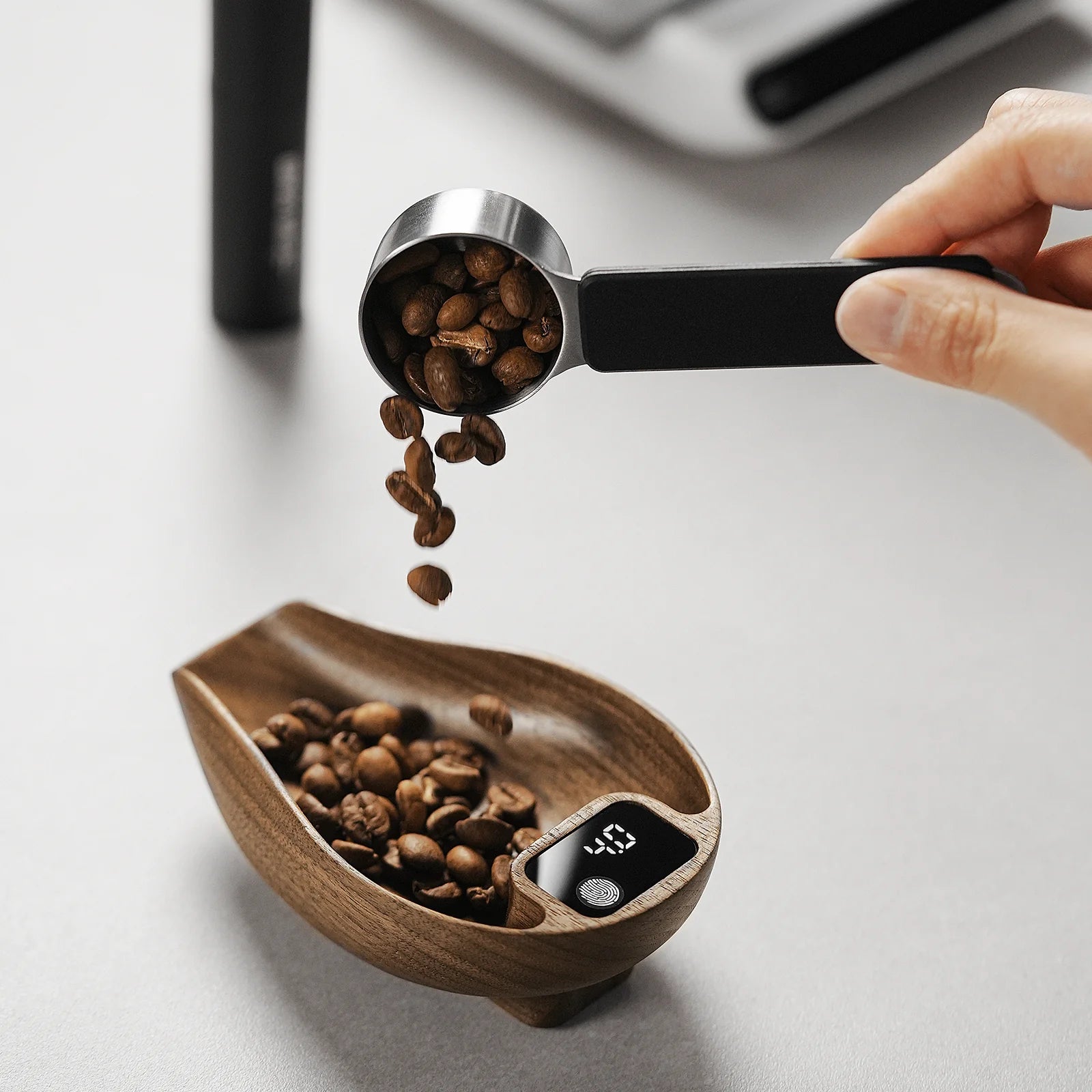 A person uses a measuring scoop to pour coffee beans into a wooden container featuring the MHW-3BOMBER Ovo Coffee Scale, which shows 45g for ideal brewing. The setup rests on a gray surface.