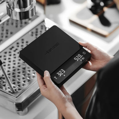 MHW-3BOMBER black coffee scale, digital display shows weight & time, held in hands. Precision brewing tool.