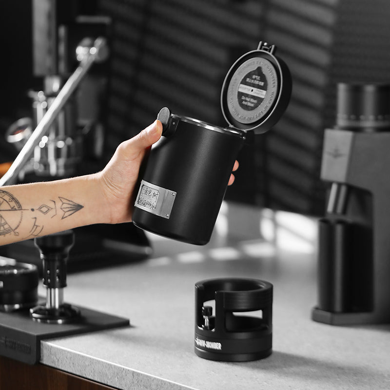 MHW-3BOMBER coffee canister with open lid. Black, features date recording. On kitchen counter, being held.