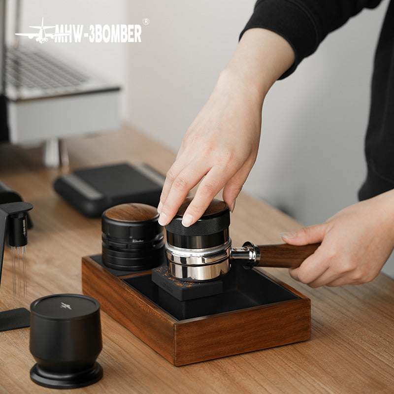 MHW-3BOMBER espresso tamping station with wood base, portafilter, tamper, and grounds container on counter.