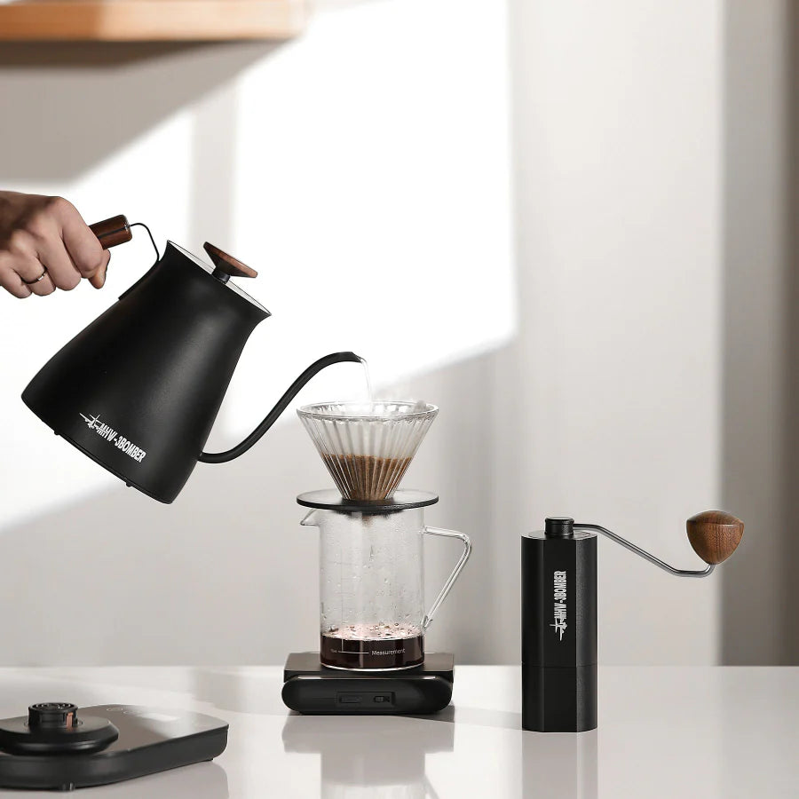 MHW-3BOMBER coffee set: Black gooseneck kettle pouring into clear pour-over, with matching hand grinder on counter.