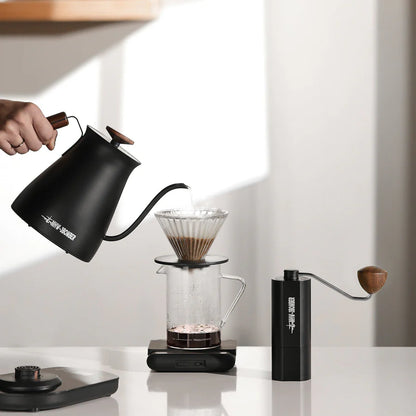 MHW-3BOMBER coffee set: Black gooseneck kettle pouring into clear pour-over, with matching hand grinder on counter.