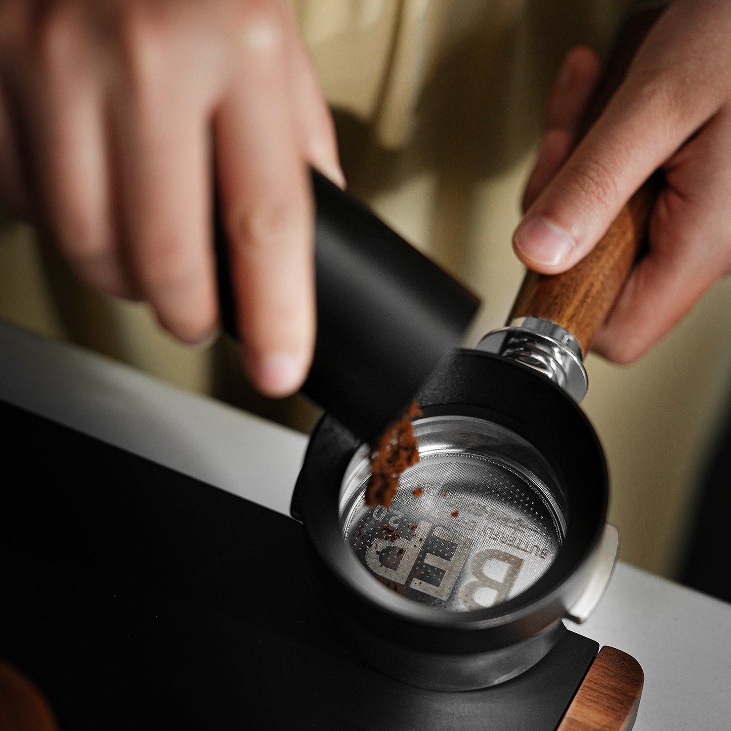 The scene shows a close-up of someone pouring ground coffee from a black container into the MHW-3BOMBER BEP Butterfly Coffee Filter Basket 2.0 with dual-layer filtration, featuring a portafilter with a wooden handle above a sleek dark surface.