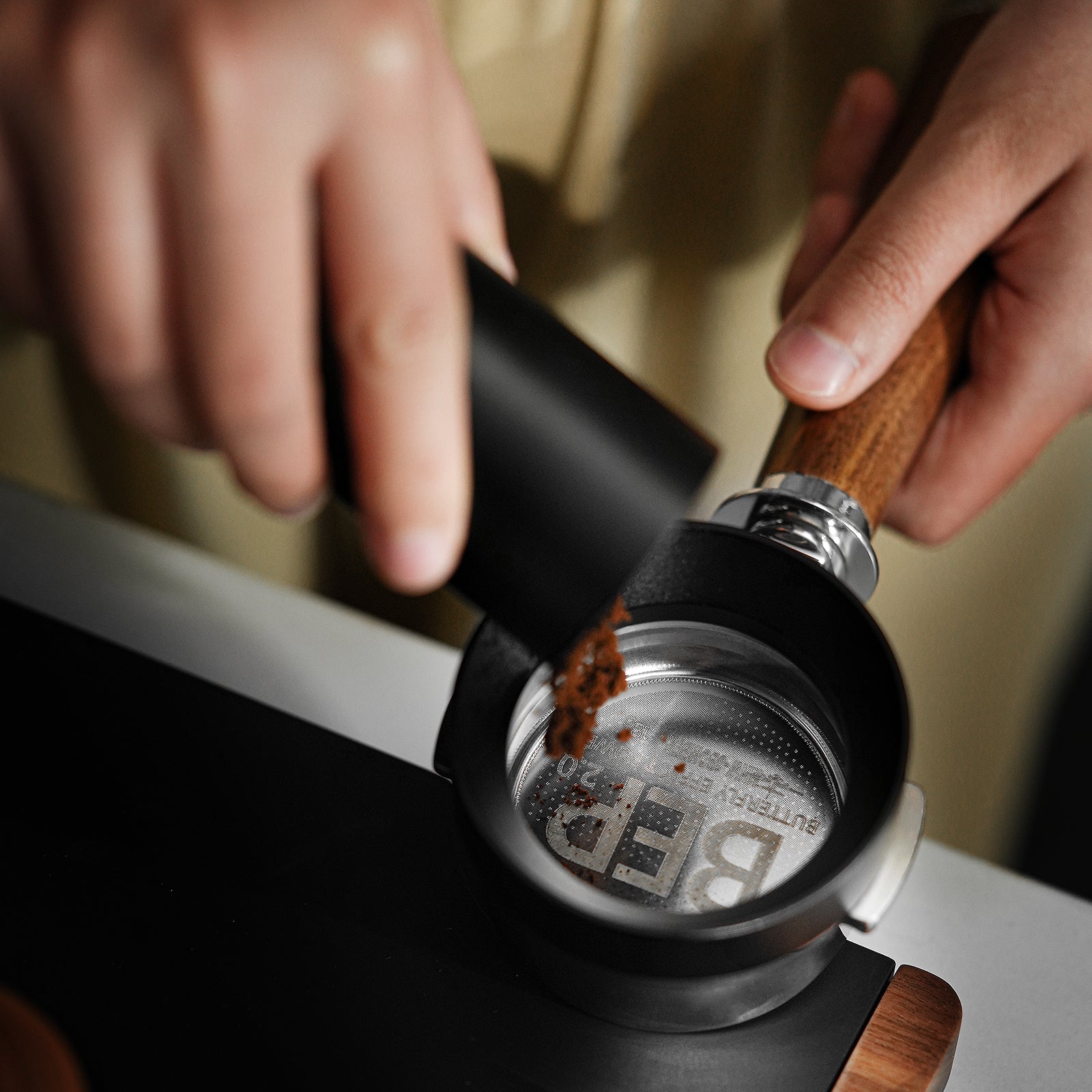The scene shows a close-up of someone pouring ground coffee from a black container into the MHW-3BOMBER BEP Butterfly Coffee Filter Basket 2.0 with dual-layer filtration, featuring a portafilter with a wooden handle above a sleek dark surface.