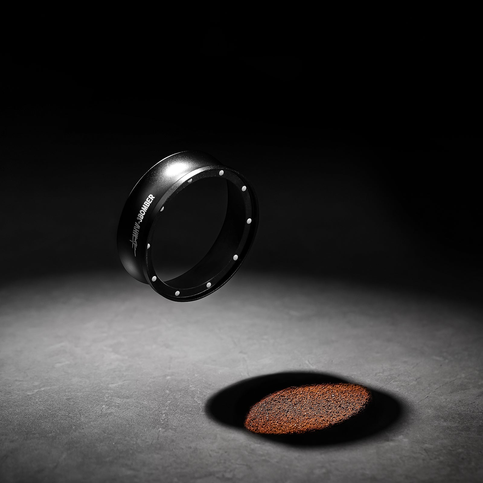 A round coffee tamper floats above finely ground coffee next to the MHW-3BOMBER Espresso Magnetic Coffee Dosing Funnel, both on a dark surface spotlighted dramatically. The tampers shadow enhances the contrast, adding to the minimalist aesthetic.