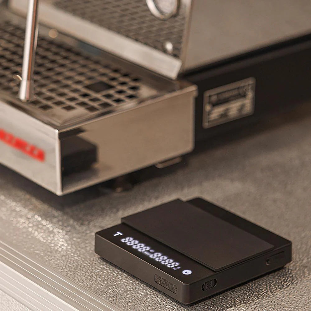 A close-up of the MHW-3BOMBER Espresso Scale Cube Coffee Scale 2.0 shows precise measurements near a coffee machine with a metal drip tray and brand logo, suggesting meticulous preparation. Its USB rechargeability enhances its modern appeal on the textured countertop.