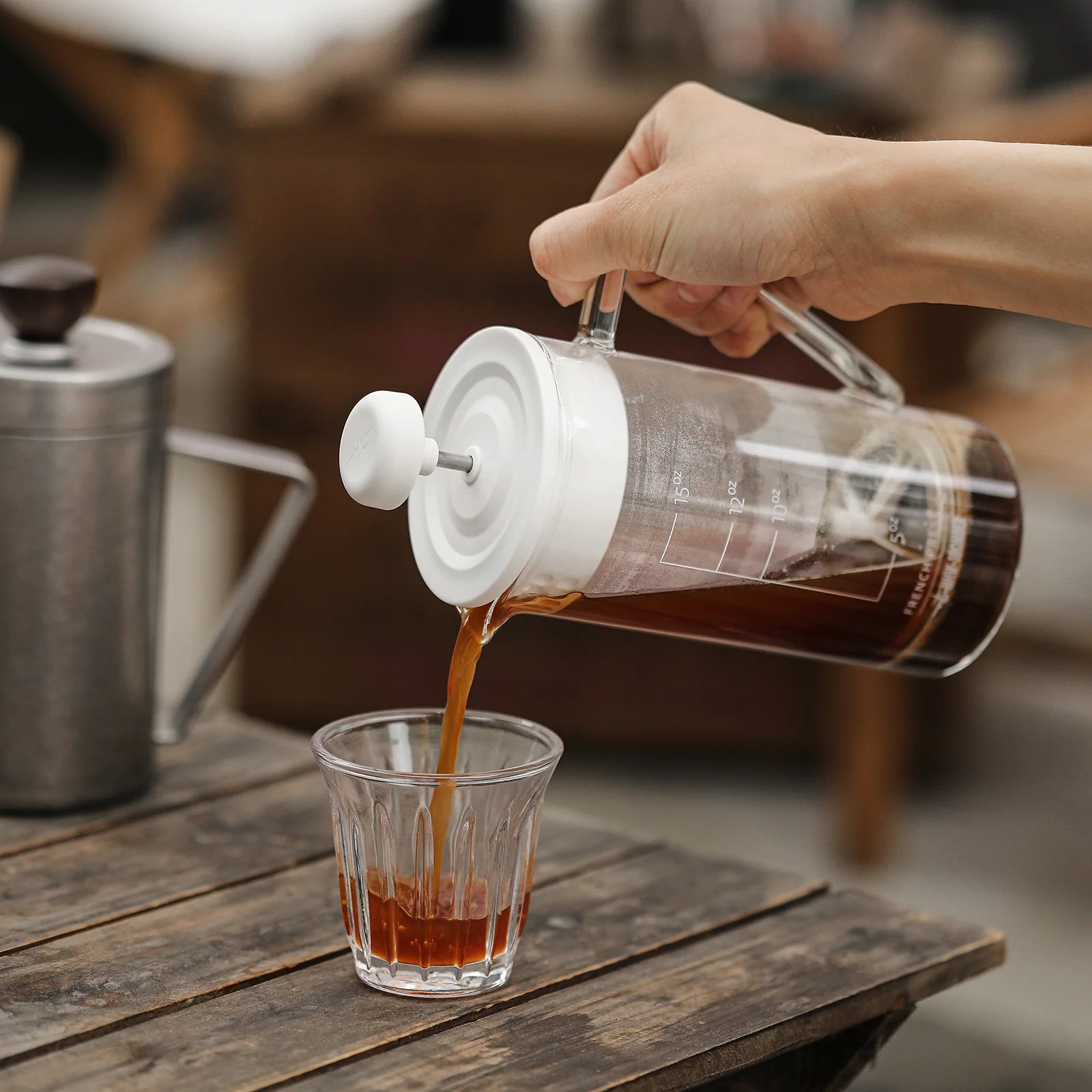 A hand pours coffee from the MHW-3BOMBER Gimme French Press, boasting a stainless steel filter into heat-resistant glass on a rustic table. A metal kettle is in the background, creating a cozy and inviting ambiance perfect for savoring fresh brew.