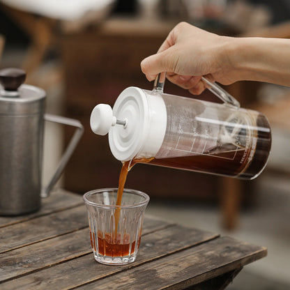 A hand pours coffee from the MHW-3BOMBER Gimme French Press, boasting a stainless steel filter into heat-resistant glass on a rustic table. A metal kettle is in the background, creating a cozy and inviting ambiance perfect for savoring fresh brew.