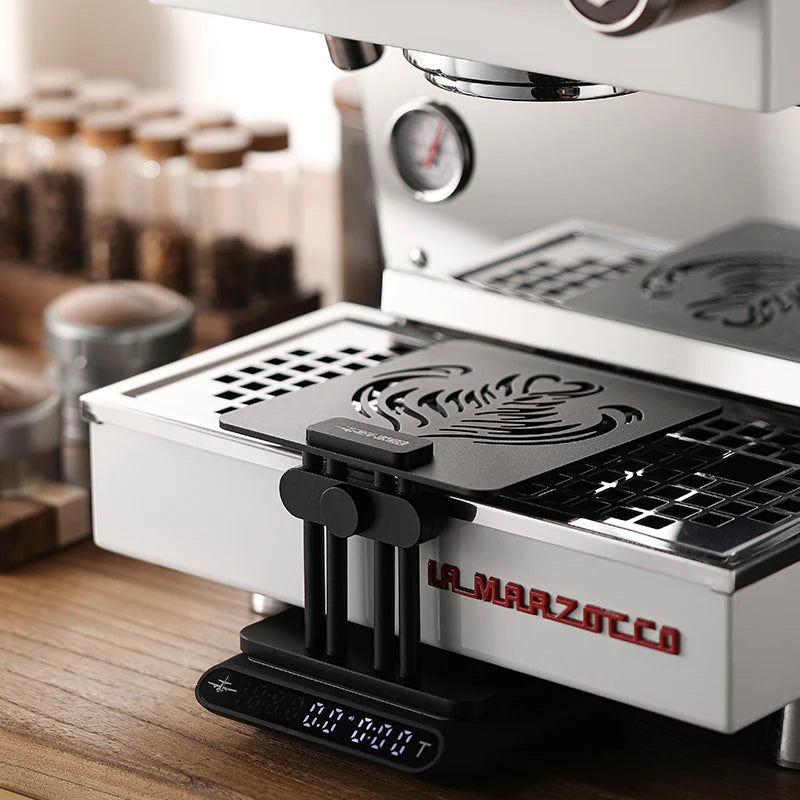 MHW-3BOMBER black coffee scale stand with adjustable height, non-slip base, next to La Marzocco espresso machine.