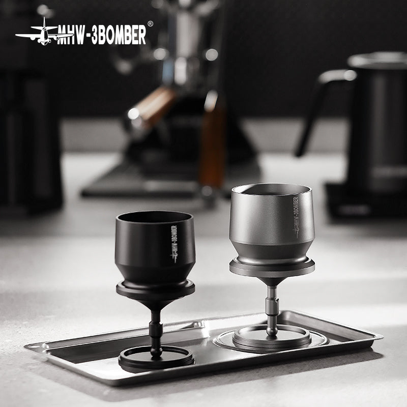 Two espresso distributors, black and silver, sit on a metal tray with the MHW-3BOMBER Espresso Coffee Powder Dosing Funnel nearby. A blurred kettle and espresso machine create a modern kitchen vibe cherished by baristas.