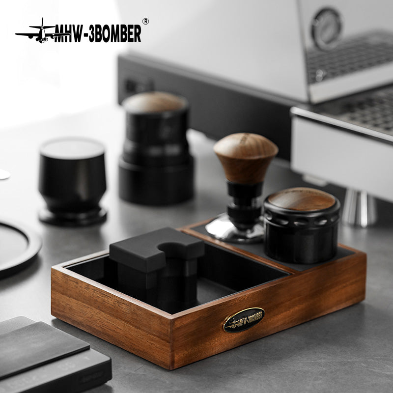 MHW-3BOMBER espresso tamping station. Wooden holder with black silicone mat, knock box, tamper, and distributor on counter.