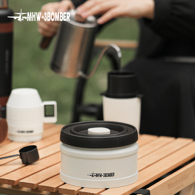 MHW-3BOMBER white vacuum coffee canister with black lid on a wooden table. Person pouring coffee in background.