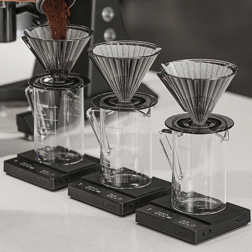Three clear beakers with black drippers sit on MHW-3BOMBER Espresso Scale Cube Coffee Scales. Coffee grounds are poured into one dripper. Each marked beaker on a light surface elegantly highlights the scales USB rechargeability.