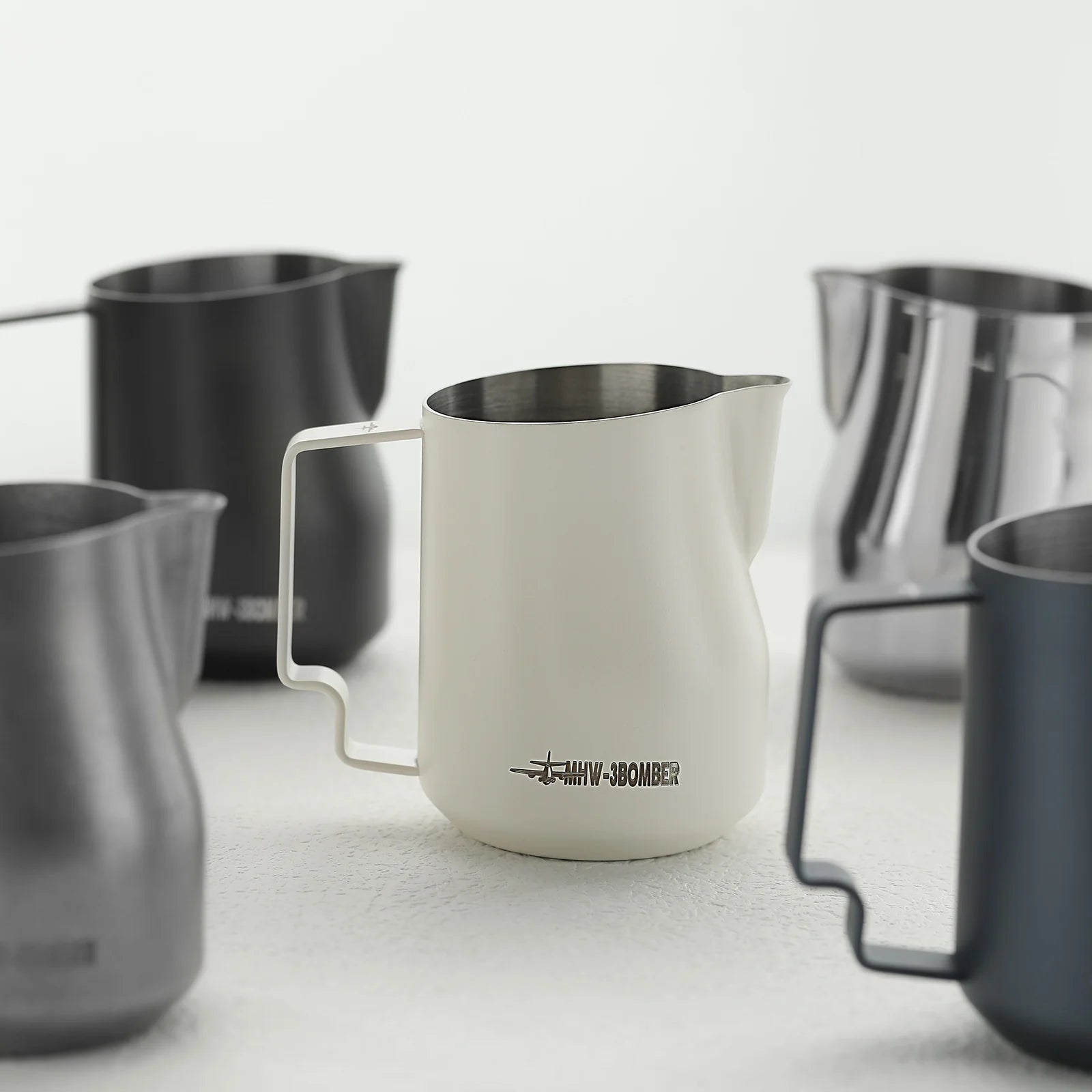 The MHW-3BOMBER Turbo Milk Pitcher collection includes five striking metallic pitchers in black, white, gray, and silver. Each stainless steel design has a handle and spout, with the branded white model perfect for latte art, elegantly displayed on a light surface.
