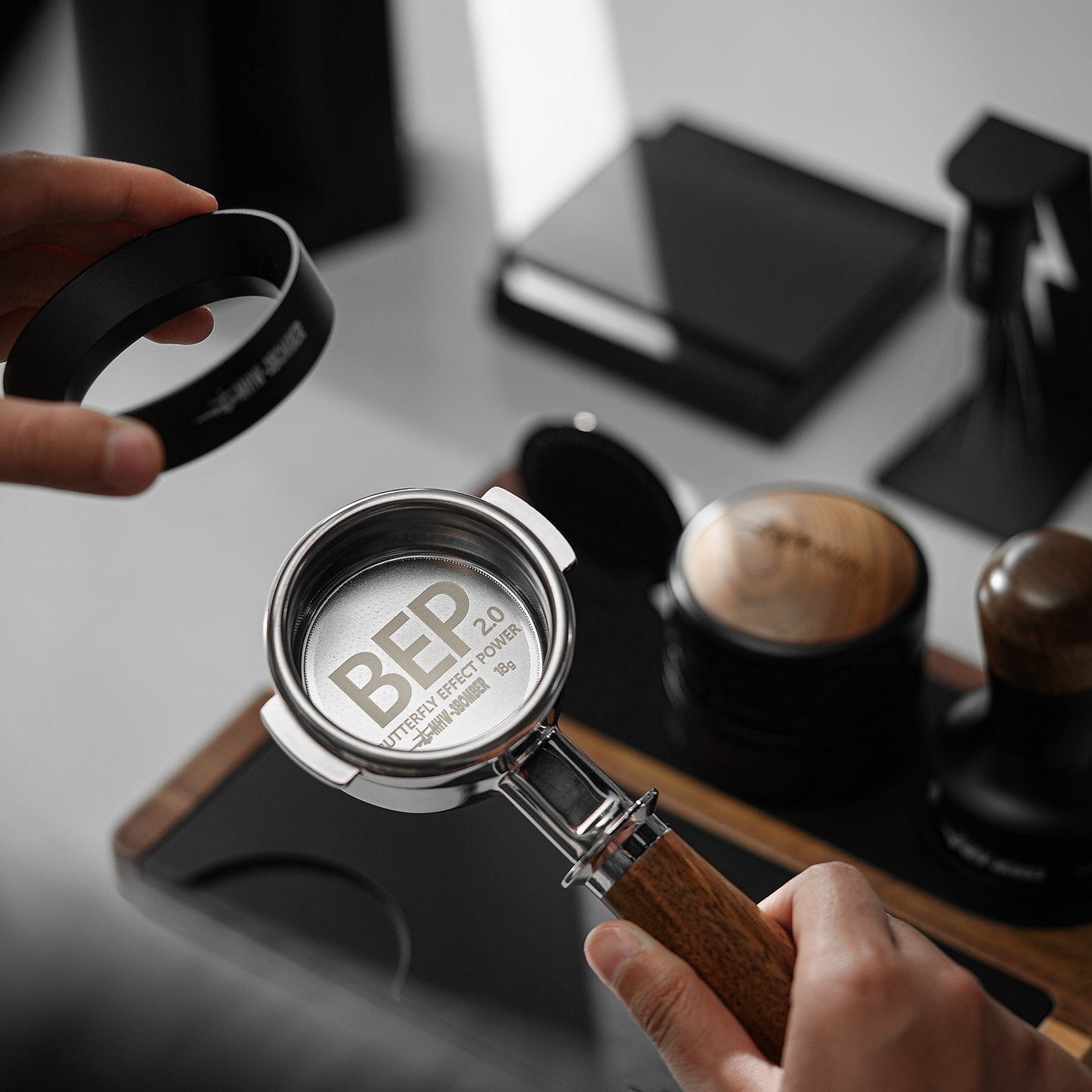 A person holds a MHW-3BOMBER BEP Butterfly Coffee Filter Basket 2.0 (58.5mm) with BEP 2.0 and Battery Effect Power text, surrounded by coffee tools like a tamper and metal ring on a sleek, modern surface, suggesting coffee preparation.