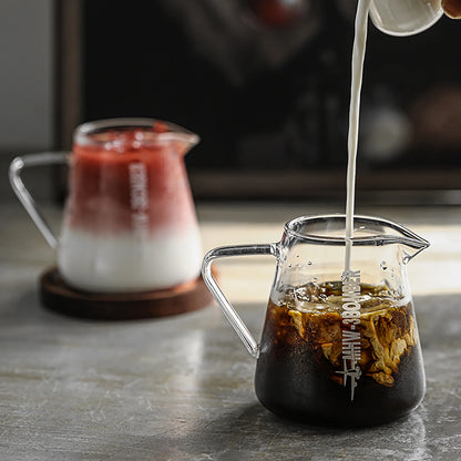 Two clear MHW-3BOMBER Elf Coffee Servers sit gracefully on the table. The front server, crafted from borosilicate glass, holds iced coffee with cream swirling in. Behind it, on a wooden coaster, another server contains a layered pink and white drink, with shadows and highlights enhancing this drip-free pour scene.