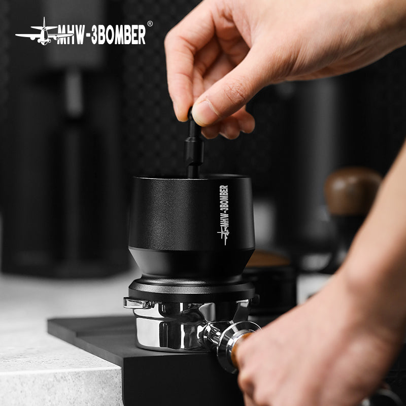 A home enthusiast expertly uses the MHW-3BOMBER espresso grinder, paired with an MHW-3BOMBER dosing funnel, to perfect their brew. The grinder rests over a portafilter on the counter, where blurred kitchen items add ambiance to their brewing experience.