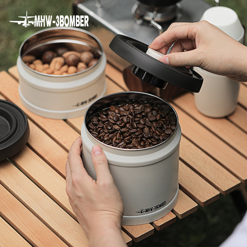 MHW-3BOMBER vacuum canister on a wooden table. Beige cylinder shape with a black lid. Contains coffee beans.