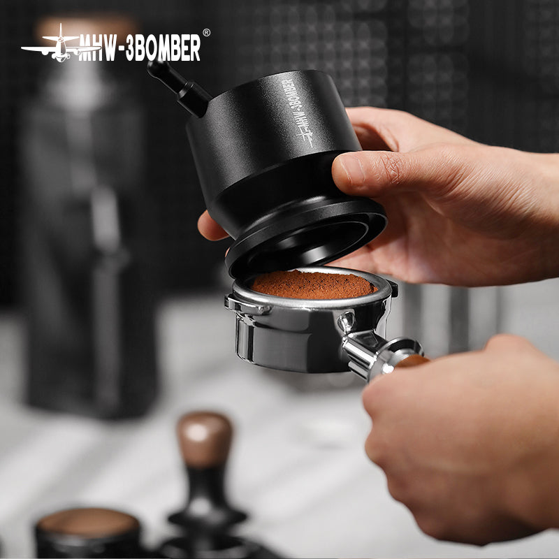 An individual, possibly a home enthusiast or barista, holds a black coffee tamper above a portafilter with tamped grounds, possibly using the MHW-3BOMBER Espresso Coffee Powder Dosing Funnel. Blurred kitchen items in the background hint at espresso brewing preparation.