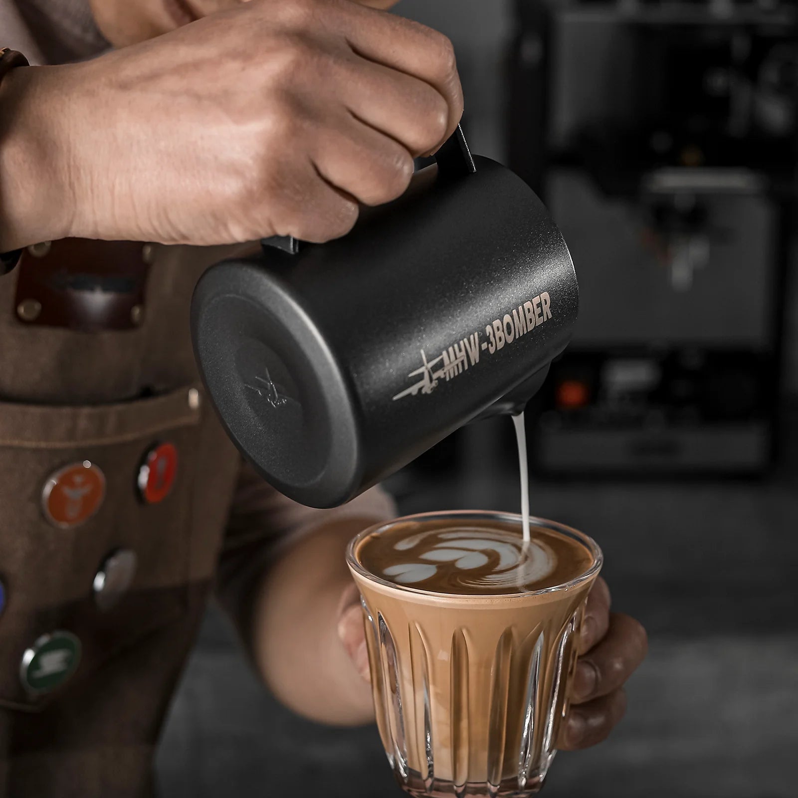 Wearing a brown apron with colorful badges, a person pours milk from the MHW-3BOMBER Latte Art Pitcher 5.0 Matte Black, featuring a crocodile spout, into coffee to create latte art against the blurred backdrop of a bustling coffee shop.