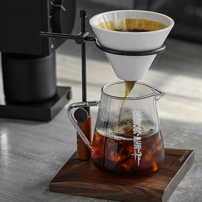 The MHW-3BOMBER Elf Coffee Server, featuring a borosilicate glass carafe on a wooden stand, uses a metal frame to hold the white cone-shaped drip coffee filter. This setup ensures a drip-free pour as freshly brewed coffee flows into the carafe atop the gray tabletop.